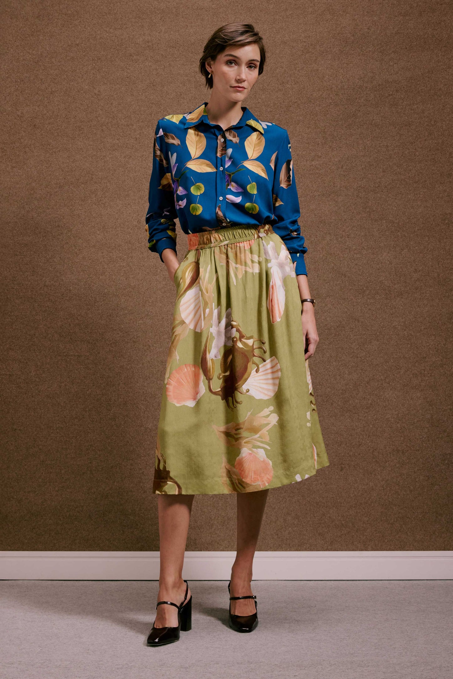 Saia Rodada Lembranças Verde midi, estampa tropical de conchas e corais em verde-oliva e laranja, com cintura elástica.