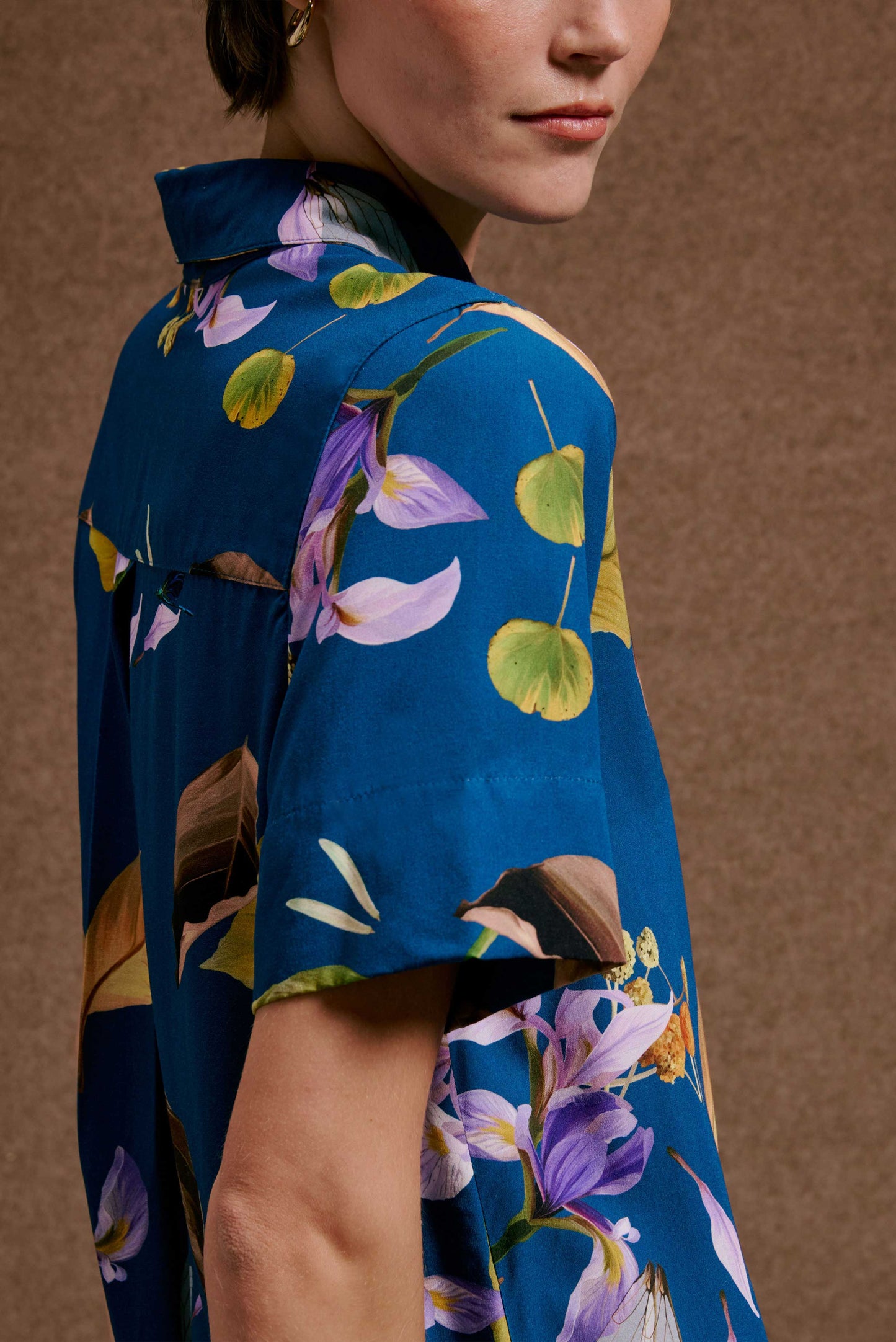 Chemise Fenda Anoitecer Botânica azul vibrante, estampa de flores roxas, lilás e folhas verdes e marrons.