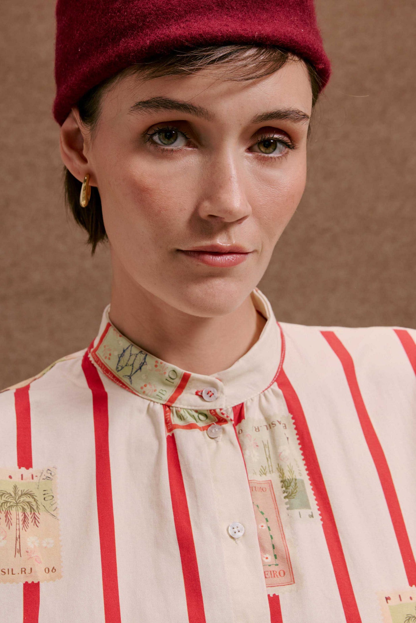 Camisa Gênova Selos Listras: Modelo veste camisa bege com listras vermelhas e estampas detalhadas de selos.