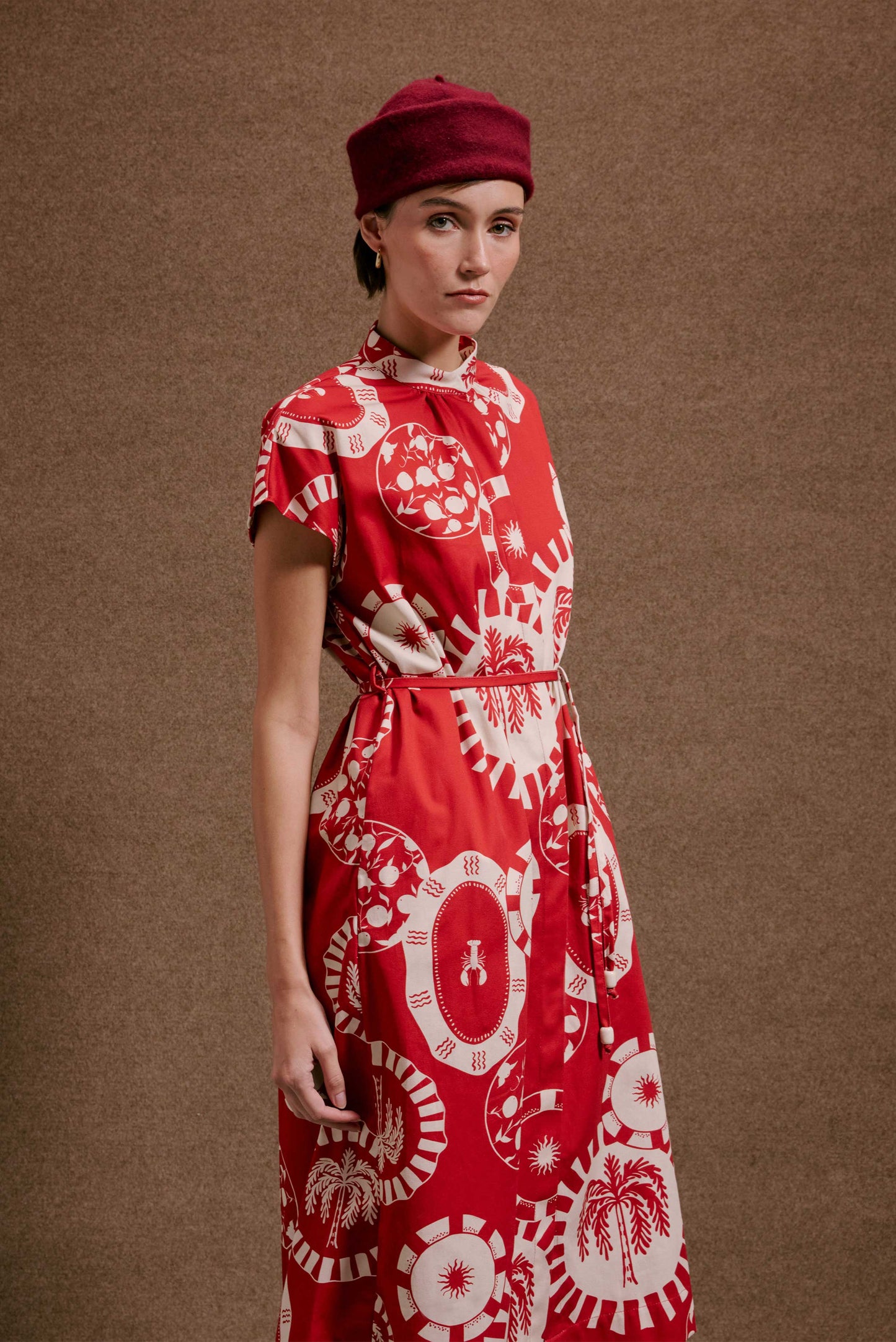 Vestido Gênova Pratinhos Vermelho, estampa branca de sol, coqueiro e lagosta, gola alta, mangas curtas e cinto.