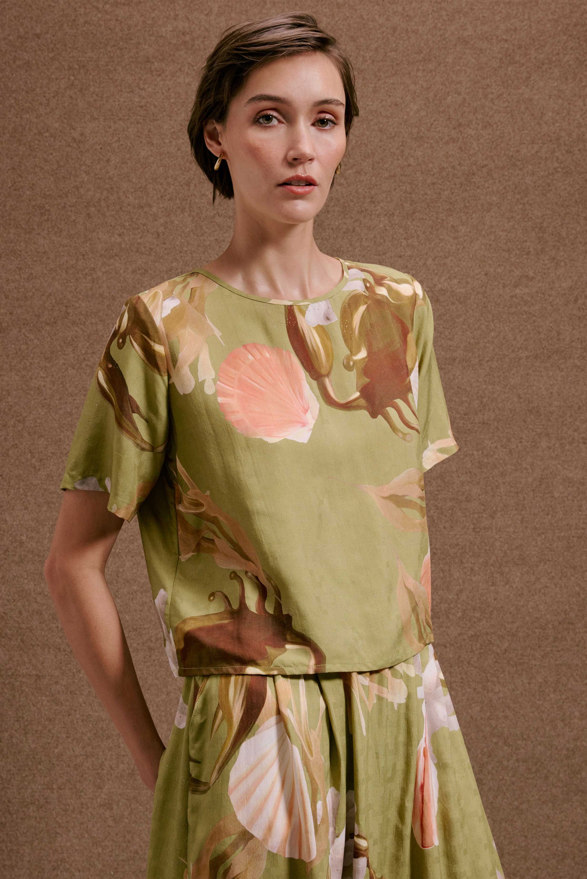 Blusa Nápoles Lembranças Verde, manga curta, estampa de conchas e folhagens verde, rosa e marrom, texturizada.