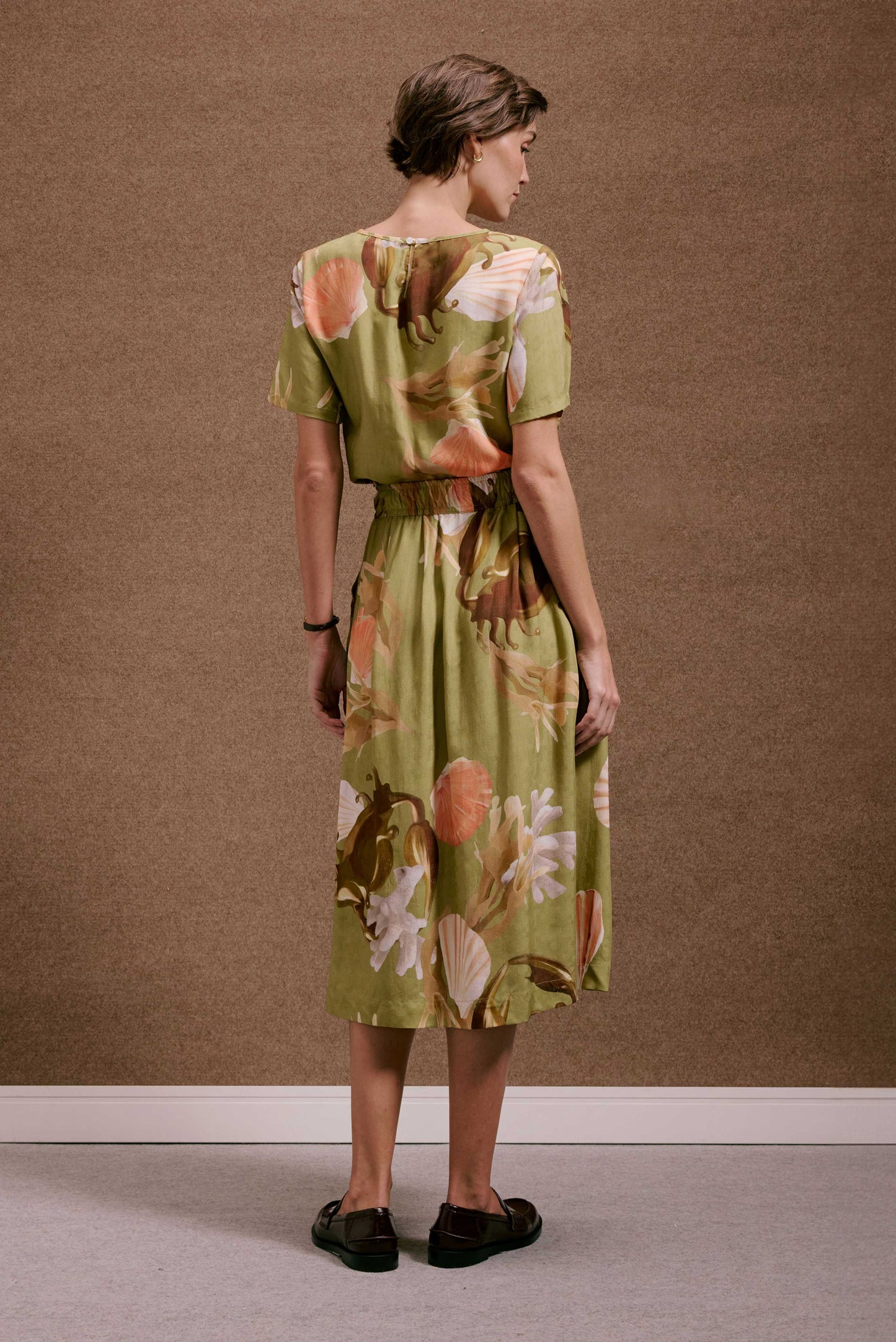 Saia Rodada Lembranças Verde com estampa de conchas e corais em tons alaranjados e terrosos, caimento leve e fluido.