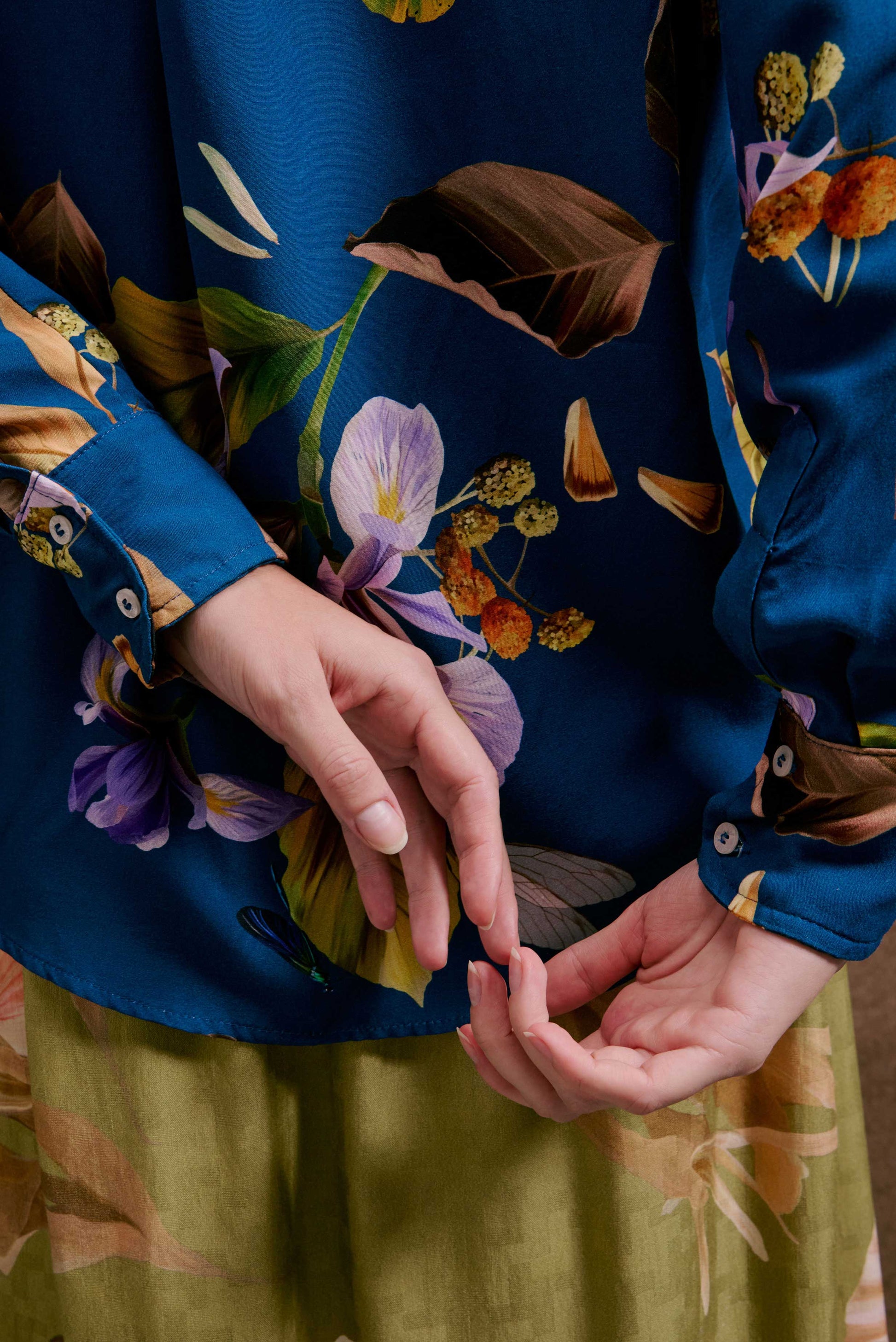 Camisa ML Anoitecer Botânica: azul-petróleo, estampa floral violeta e folhas marrons. Tecido sedoso. Mãos em saia verde ol...