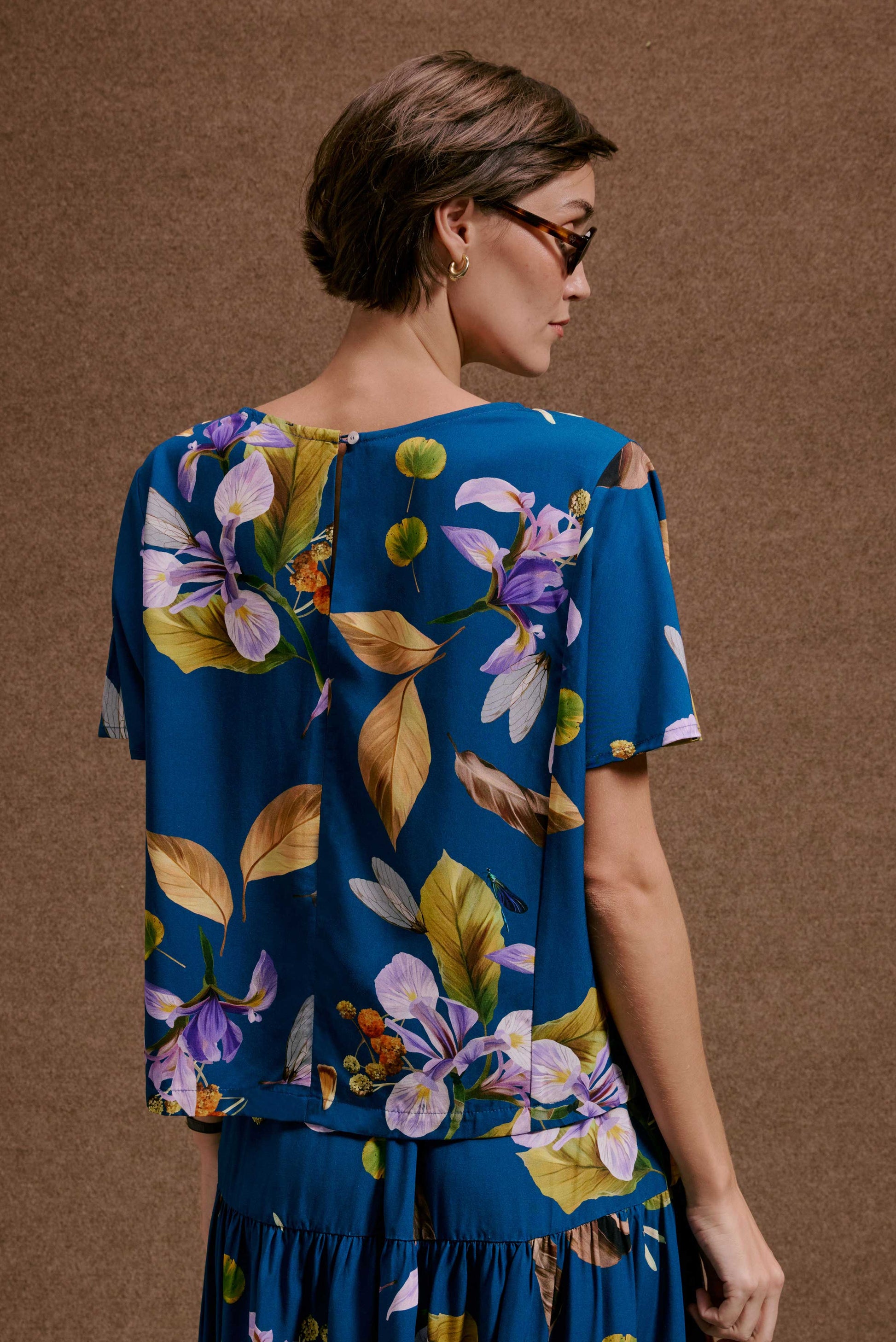 Blusa Japa New Anoitecer Botânica azul, com estampa vívida de lírios roxos, folhas e libélulas. Manga curta e decote canoa.