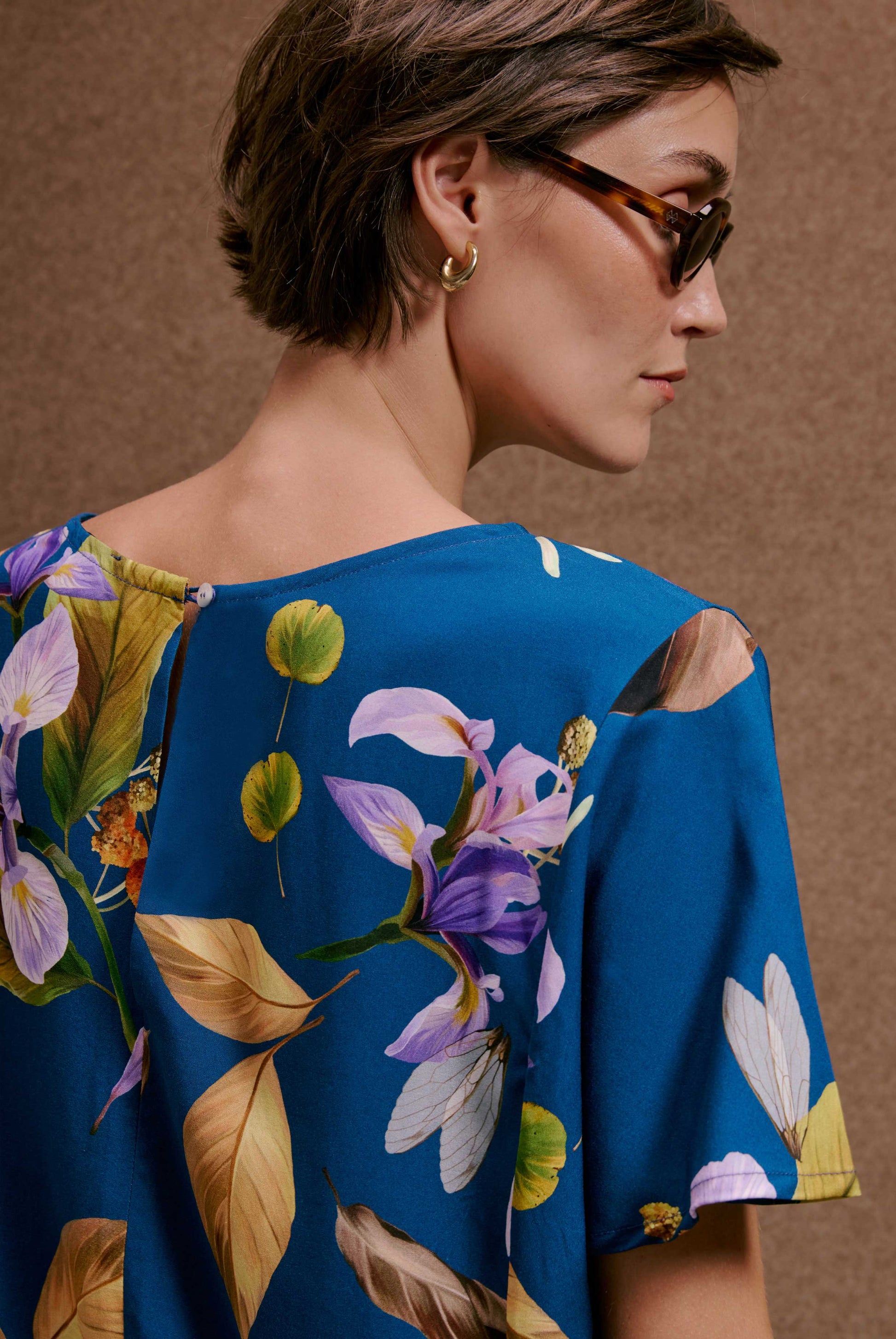 Blusa Japa Anoitecer Botânica, azul-petróleo com estampa de flores roxas, asas e folhas em tecido fluido.