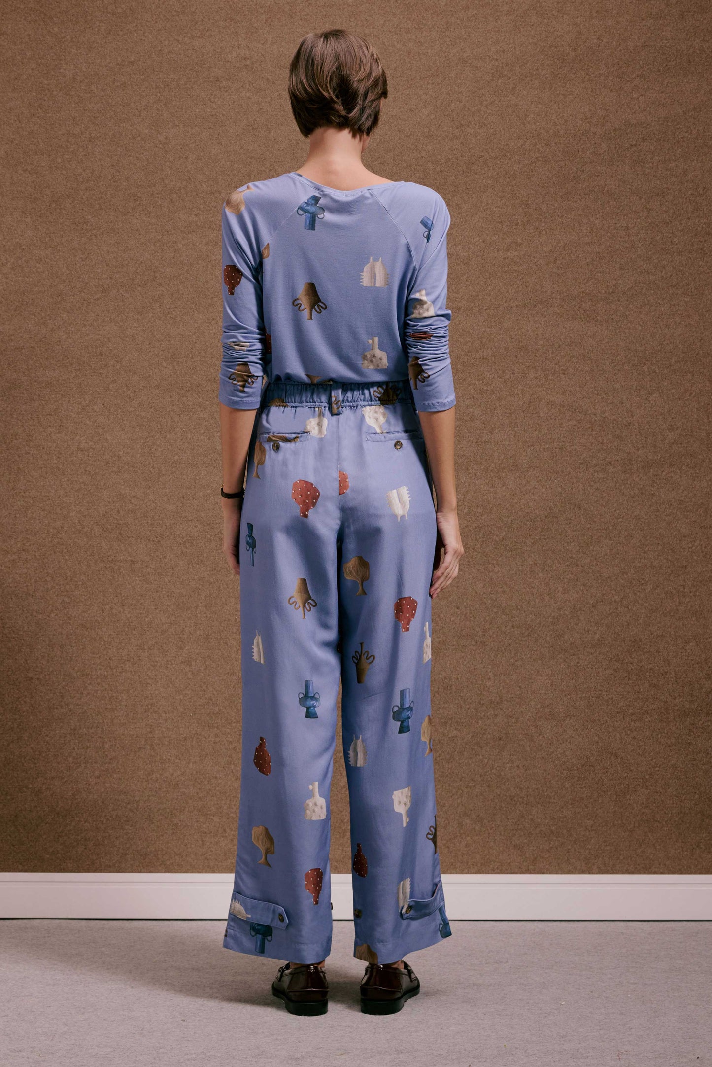Calça Porto Vasinhos Mini e blusa azul acinzentado com estampa de vasos e formas abstratas em tons terrosos.