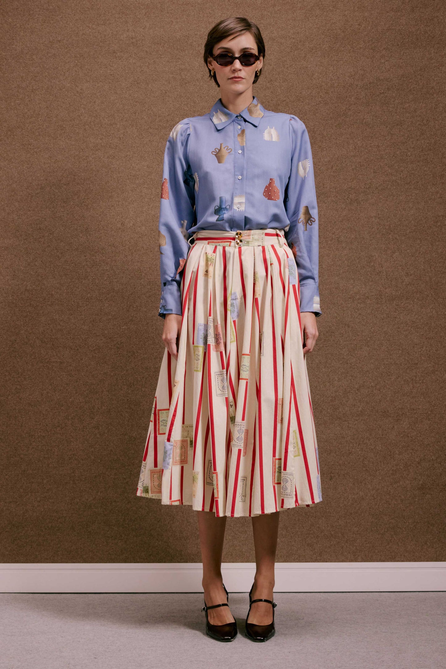 Saia Relíquia Selos Listras plissada branca com listras vermelhas e estampas de selos. Blusa azul estampada e óculos de sol.