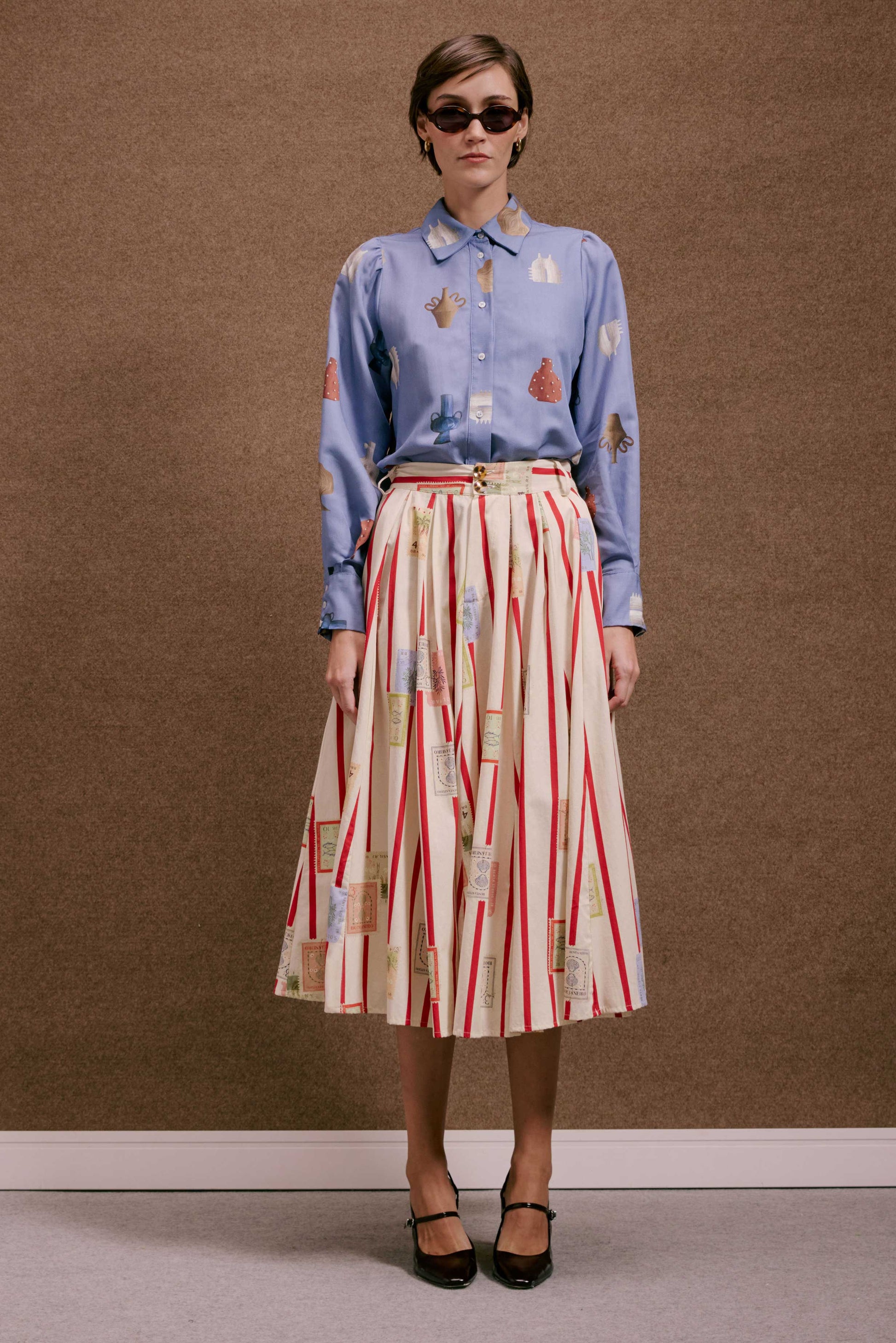 Saia Relíquia Selos Listras plissada branca com listras vermelhas e estampas de selos. Blusa azul estampada e óculos de sol.