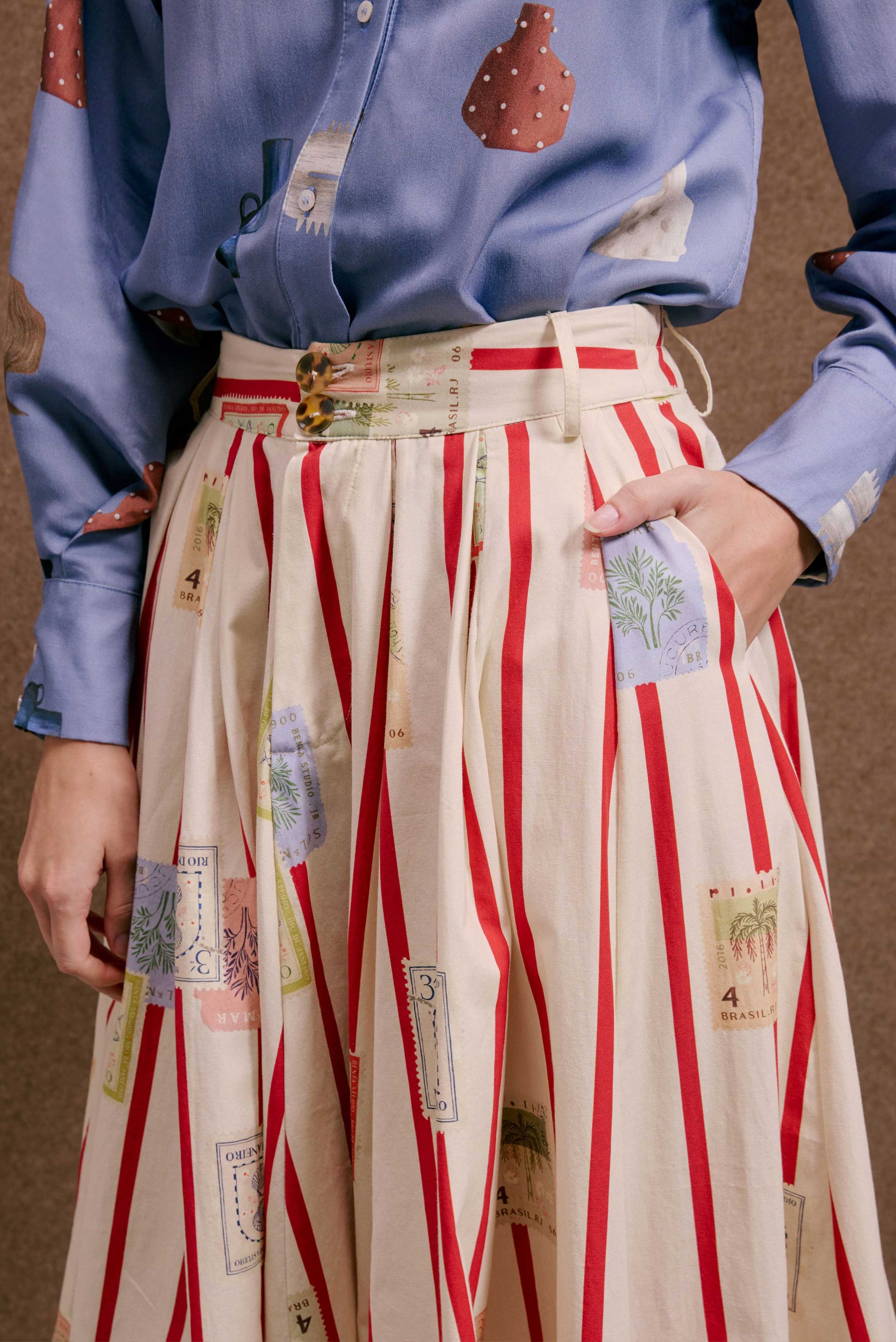 Saia Relíquia Selos Listras off-white de tecido leve com listras vermelhas e estampa de selos. Blusa azul com padrões.
