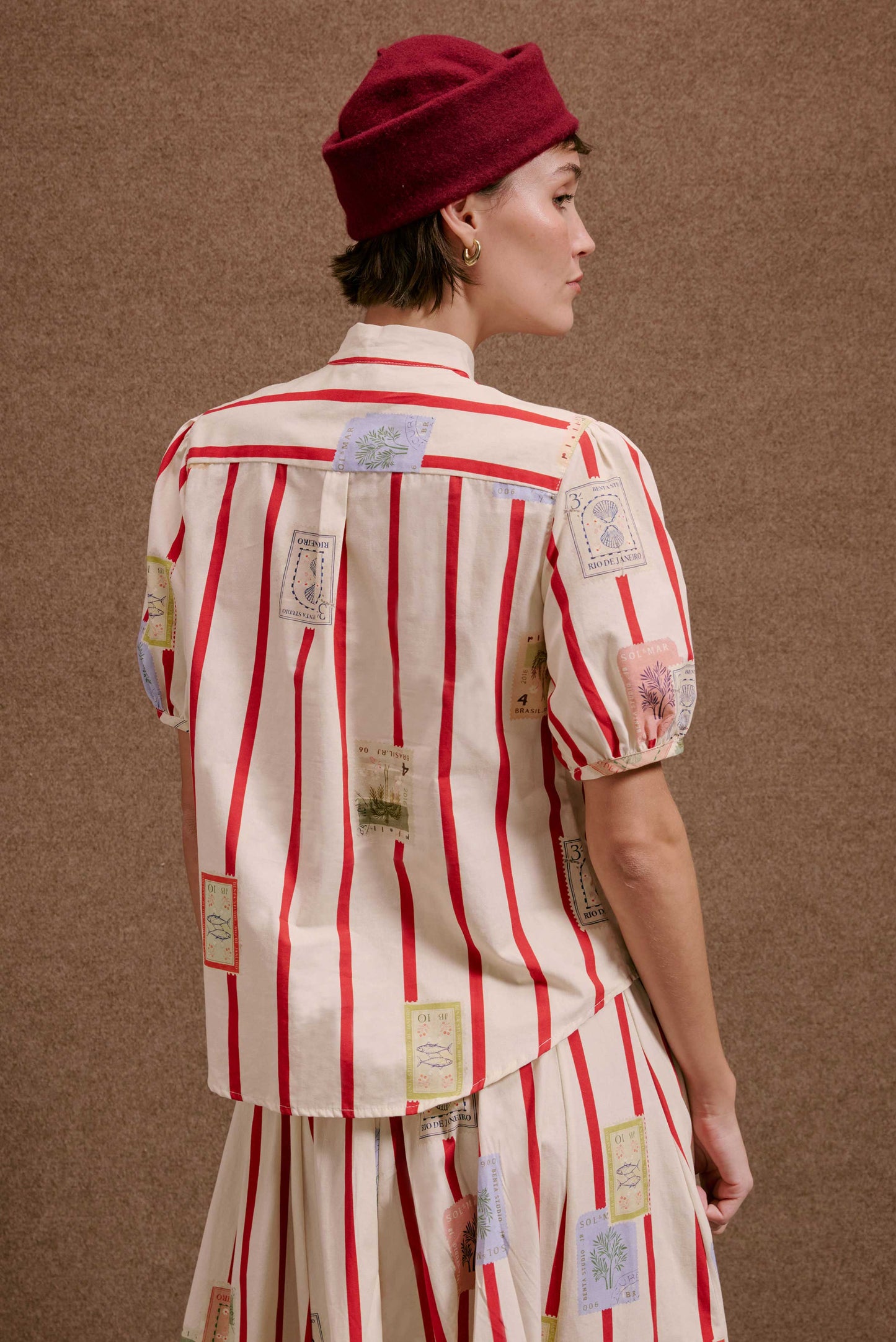 Blusa Roma Selos Listras, creme com listras vermelhas verticais e estampas de selos. Modelo com chapéu vinho.