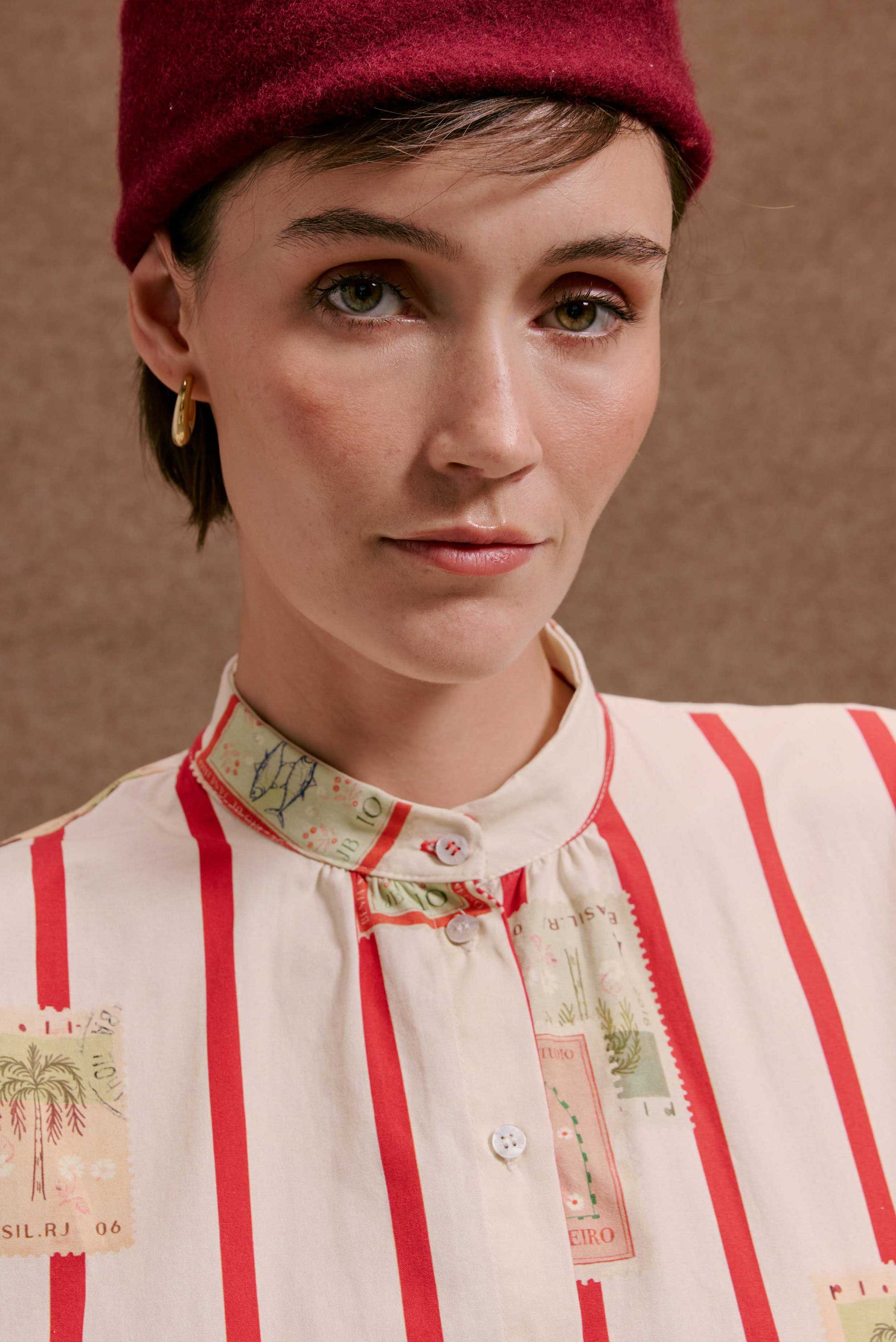 Camisa Gênova Selos Listras: Modelo veste camisa bege com listras vermelhas e estampas detalhadas de selos.