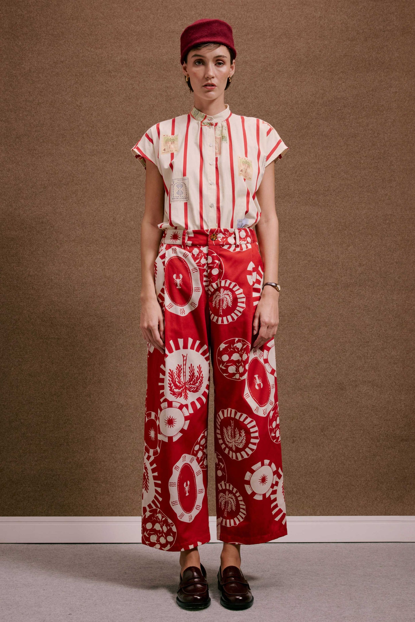 Calça Japão Pratinhos Vermelho, estampa branca com lagostas e palmeiras. Modelo usa camisa listrada vermelha e chapéu.