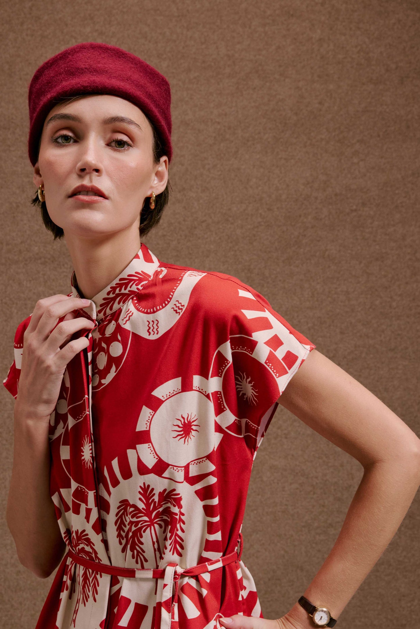 Vestido Gênova Pratinhos Vermelho com estampa de sol, palmeiras e círculos. Mulher usa boina e brincos dourados.