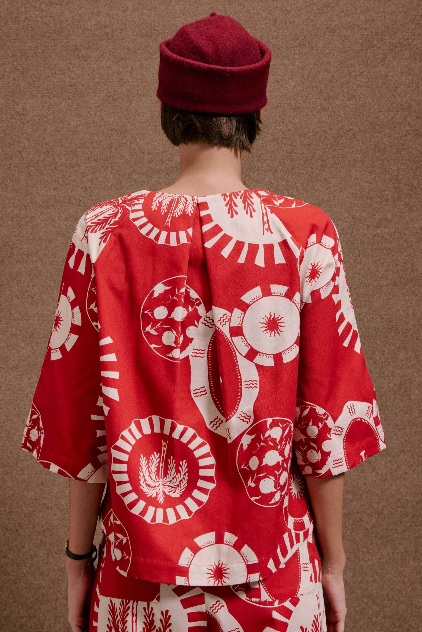 Blusa Diário Pratinhos Vermelho e branca, estampa circular com sol, coqueiros e frutas. Modelo de costas com chapéu.
