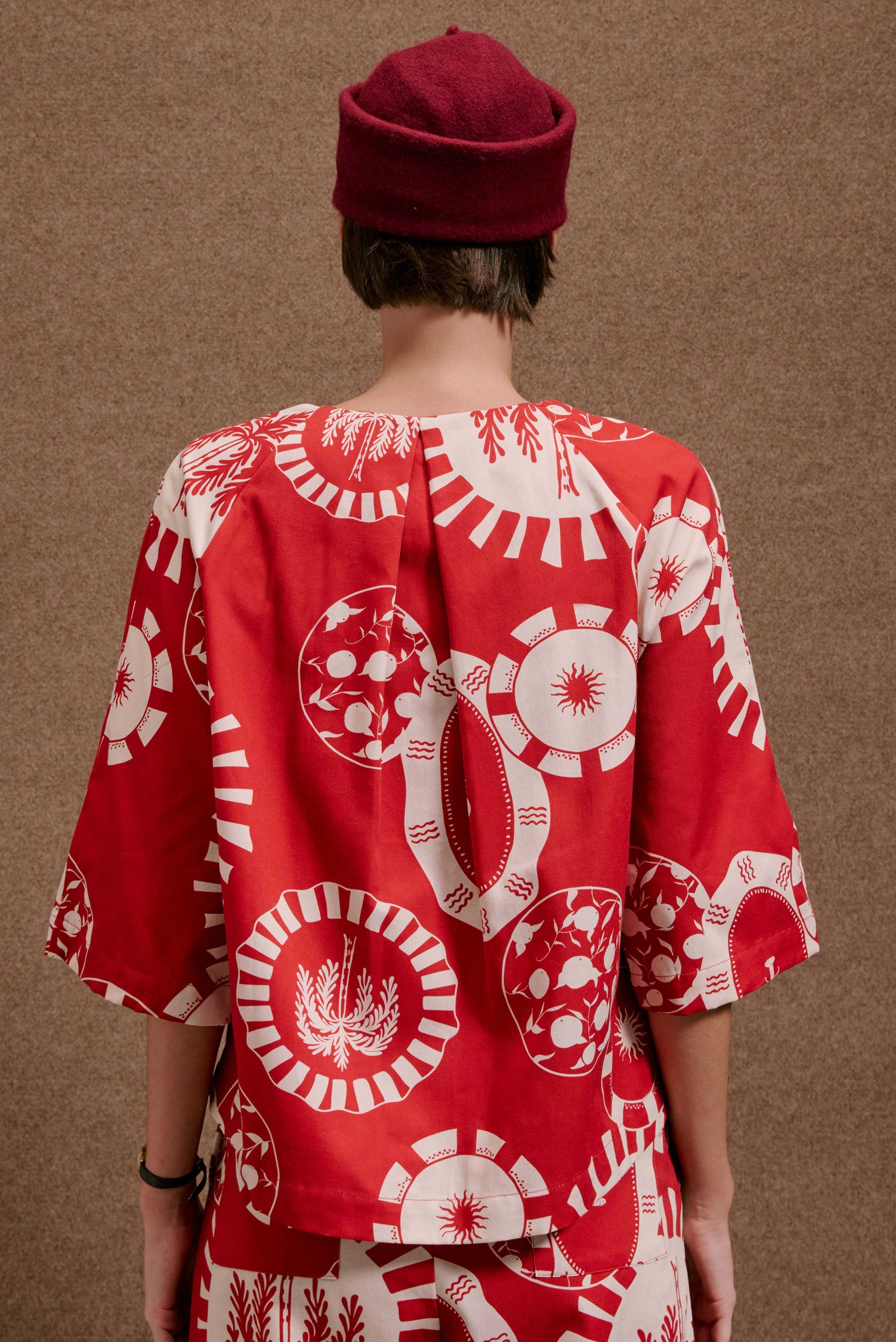 Blusa Diário Pratinhos Vermelho e branca, estampa circular com sol, coqueiros e frutas. Modelo de costas com chapéu.