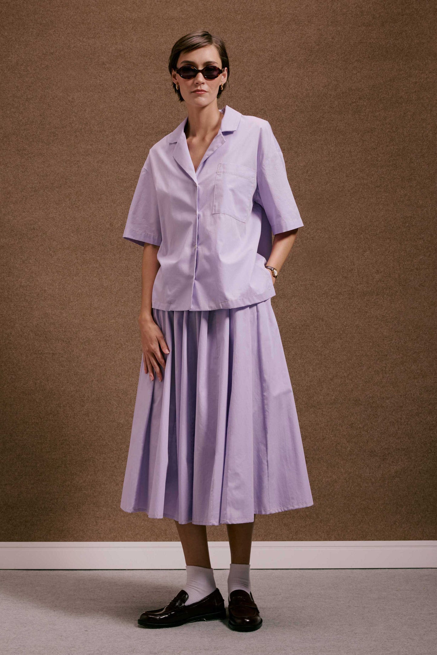 Modelo veste Camisa Havana Orquidário lilás de algodão e saia midi plissada. Look casual monocromático com loafers e óculos.
