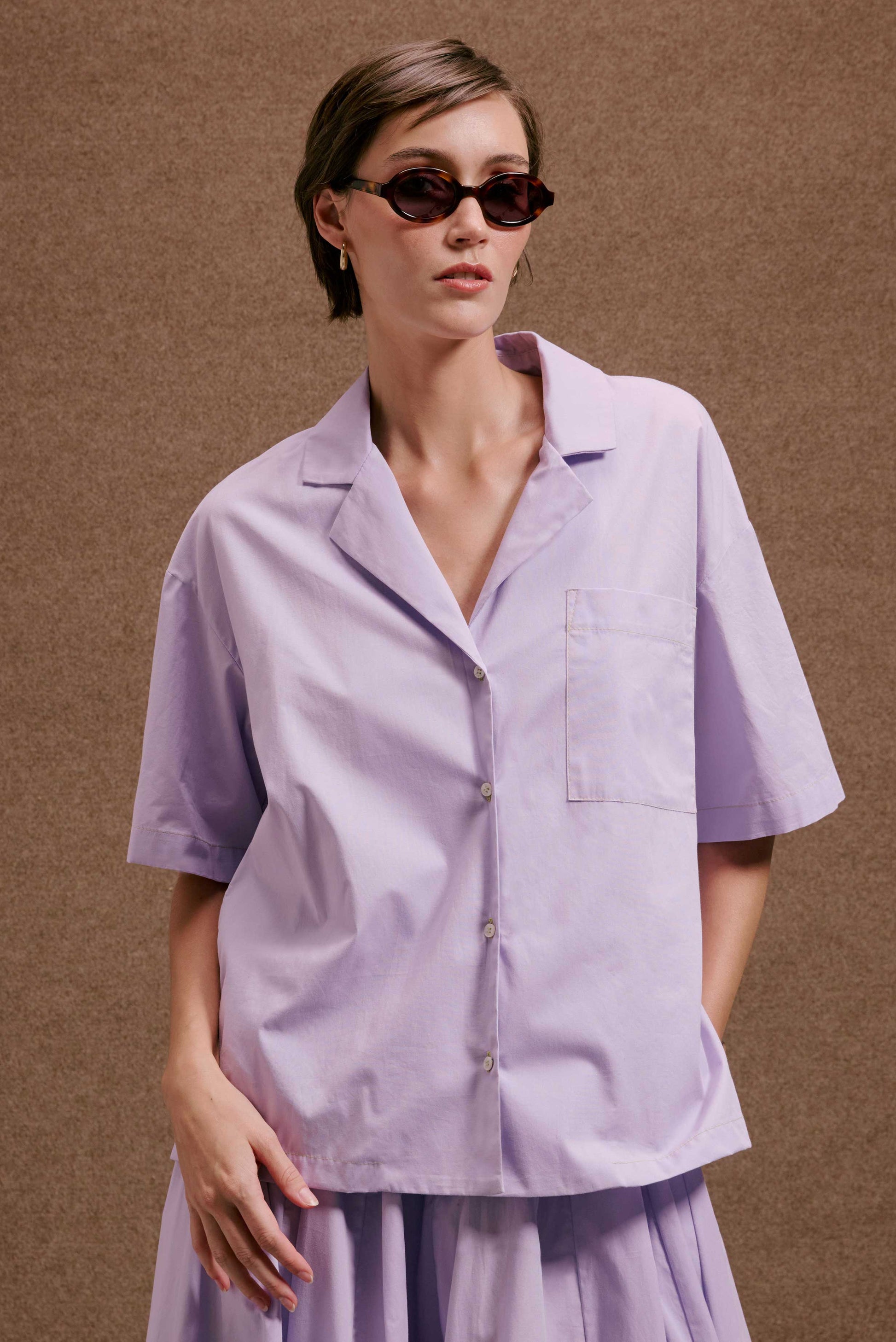 Camisa Havana Orquidário lilás, manga curta, gola lapela. Conjunto com saia plissada. Modelo com óculos de sol.