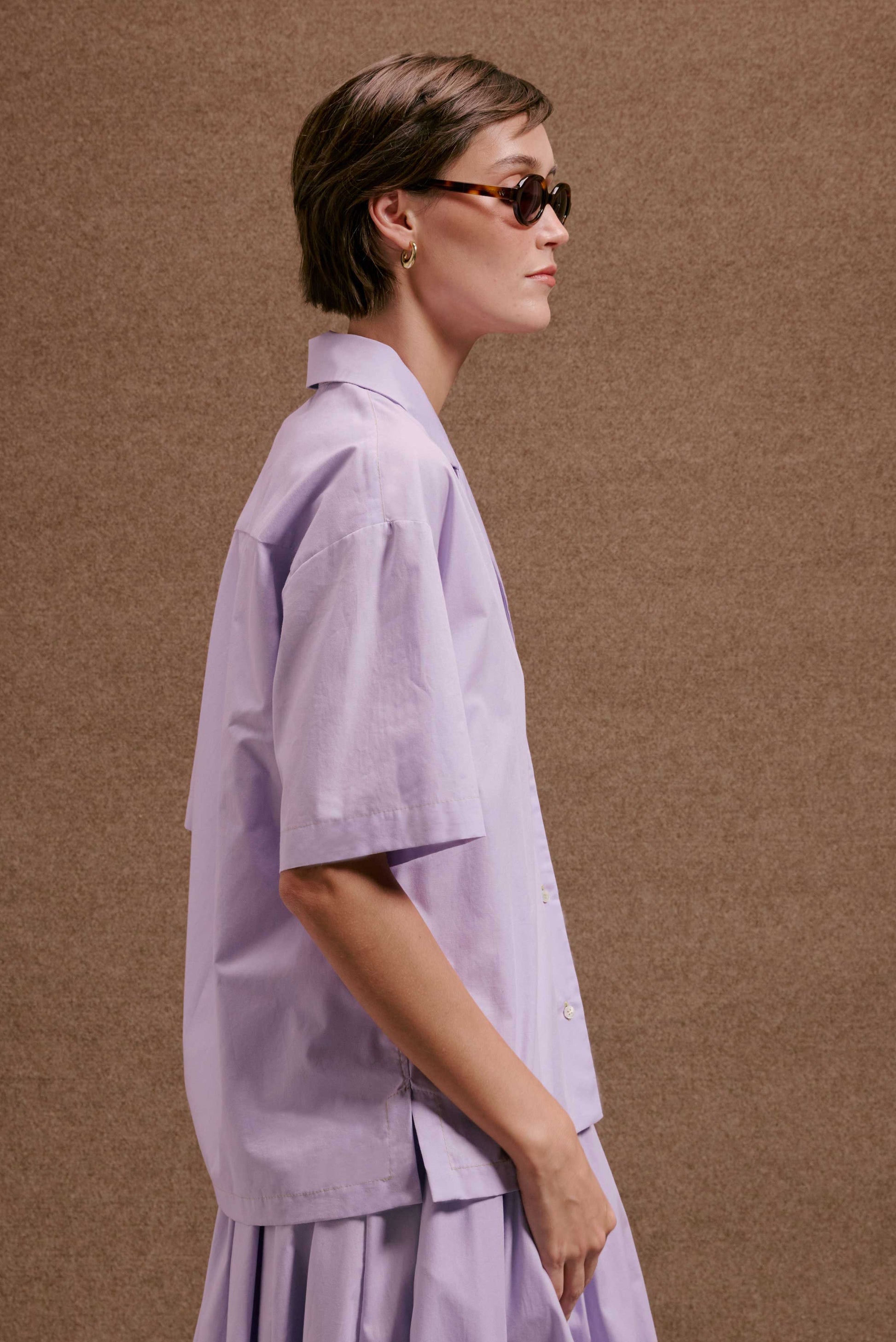 Mulher de perfil usando Camisa Havana Orquidário lilás de manga curta com saia. Óculos e brinco dourado.