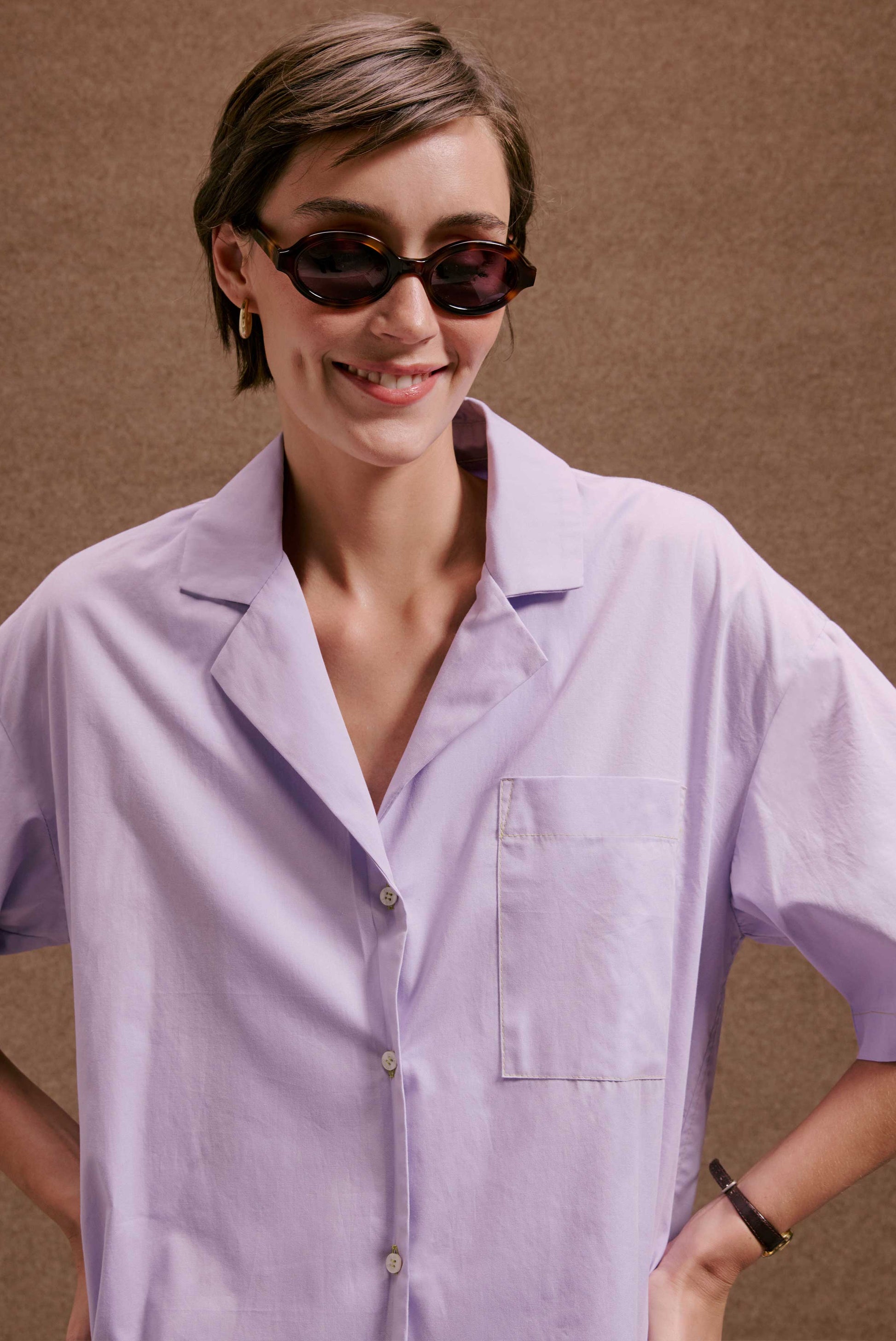 Camisa Havana Orquidário lilás, manga curta, bolso e botões, tecido leve. Mulher sorrindo de óculos escuros.